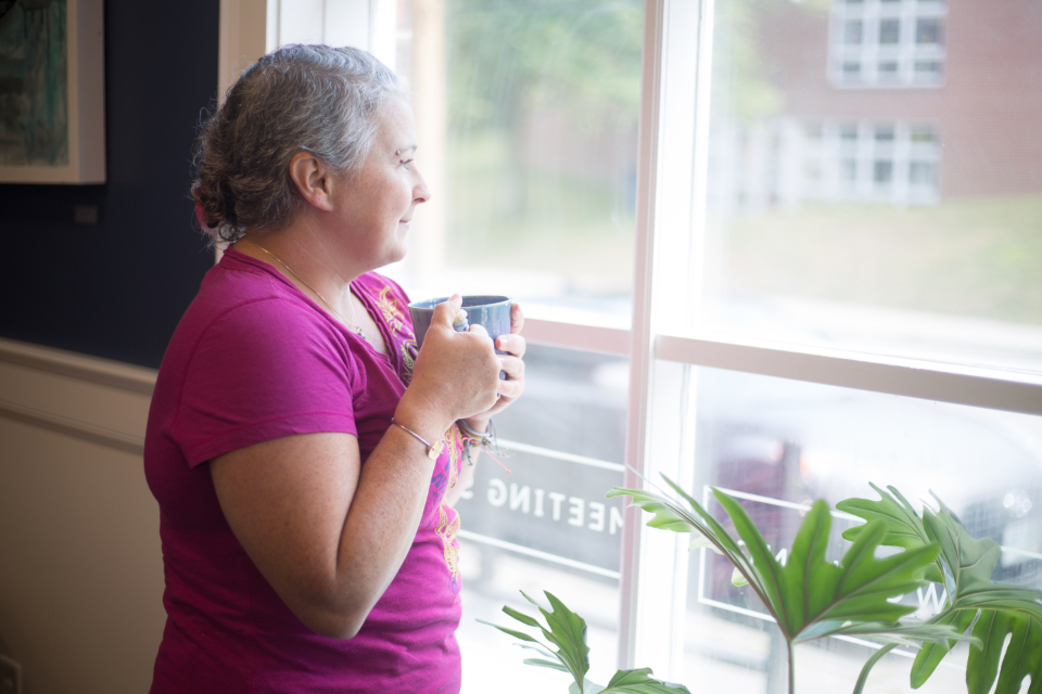 Mature woman holding a warm drink and looking out of a sunlit window