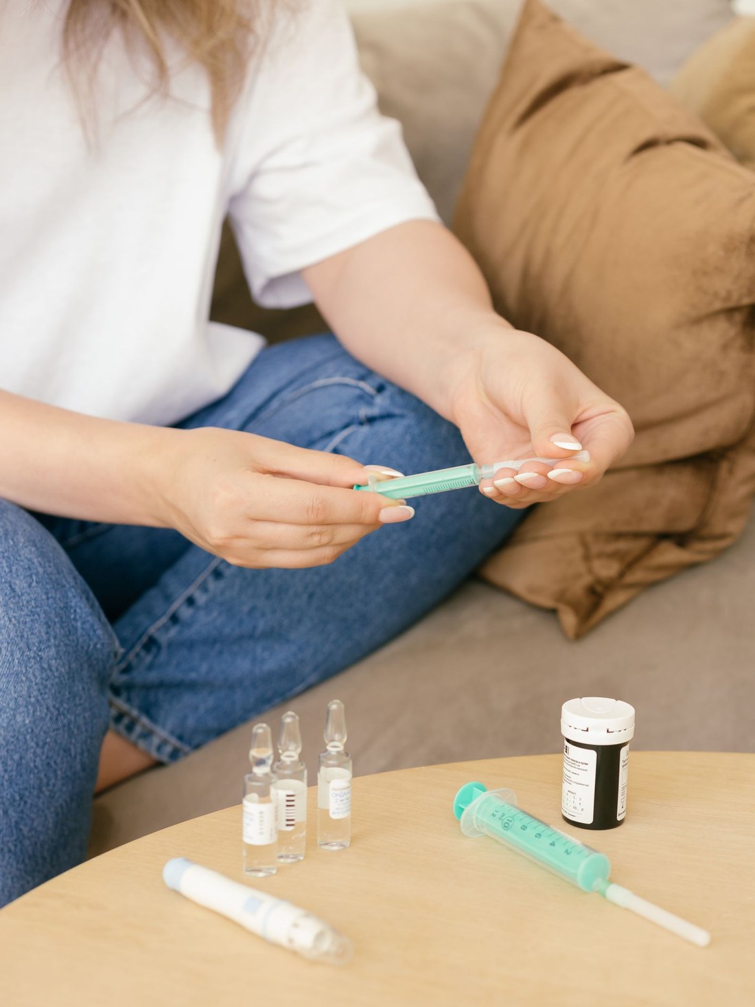 A woman sitting on a couch preparing a syringe, with ampoules and a medication bottle on a wooden table